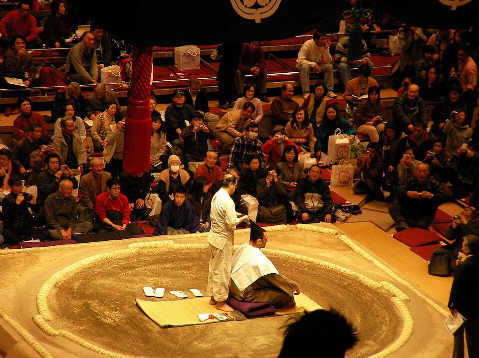 A wrestler kneeling in the ring while a man in white cuts his topknot before a full arena.